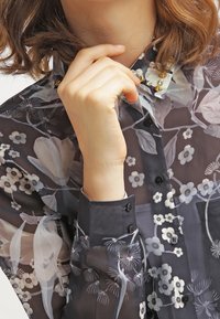 Transparente Bluse mit Blumenmuster in dunklen Tönen, mit einem Knopfverschluss, detaillierten Blumenmustern und einem verzierten Kragen mit Akzenten.