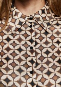 Hemd aus durchscheinendem Stoff mit geometrischem Muster aus Kreisen und Sternen in Braun, Creme und Schwarz, mit schwarzen Knöpfen.