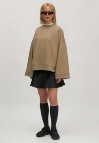 Vrouw in een beige oversized sweatshirt, donkere flared rok, kniehoge sokken, zwarte schoenen, zonnebril en gouden oorbellen tegen een effen achtergrond.