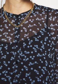Blouse noire transparente avec petit imprimé floral bleu, boutons noirs et collier chaîne dorée autour du cou et du haut de la poitrine d’une femme.