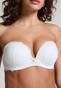 Vit spets-bh utan axelband med blommönster, djup urringning, vågiga kanter och en dekorativ rosett med en liten ädelsten i mitten.