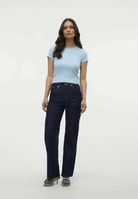 Gaiši zila īsām piedurknēm blūze, tumši zili taisnas kājas džinsi un pelēkas augstpapēžu kurpes, stāvot uz vienkārša baltā fona.