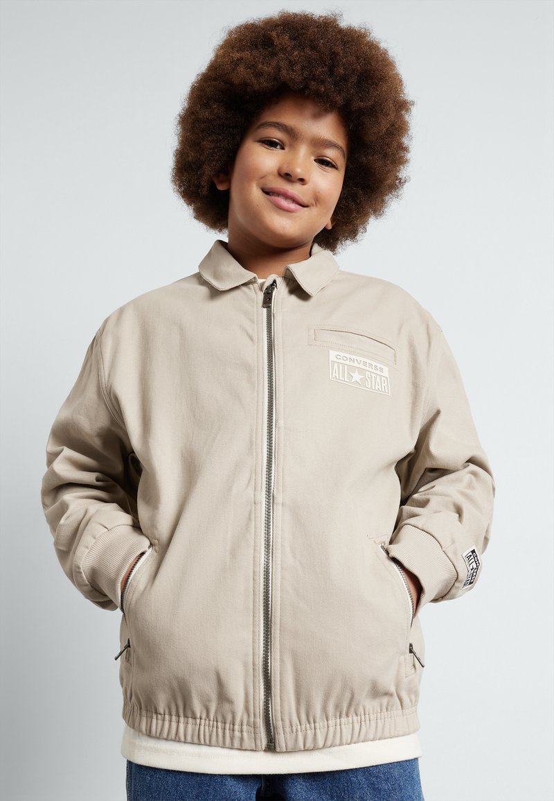 Beige jacka med dragkedja fram, ribbad mudd och elastisk nederkant; har 'Converse All Star'-logotypen på bröstfickan.