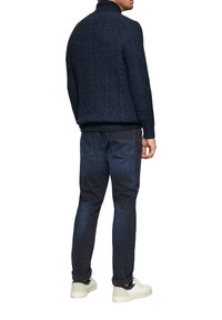 Pull en maille bleu marine avec un motif de losanges texturé, col montant et poignets côtelés, associé à un jean en denim foncé et des baskets blanches.