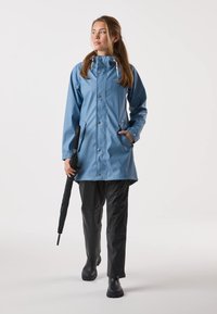 Blå vattentät regnrock med huva, tryckknappar, två sidofickor, tillsammans med svarta vattentäta byxor och stövlar, håller ett svart paraply.