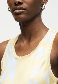 Κοντινό πλάνο ενός ατόμου που φορά χρυσούς κρίκους στα αυτιά και ένα αμάνικο τοπ με κίτρινο και ανοιχτό μπλε σχέδιο, με φόντο ένα μονόχρωμο υπόβαθρο.