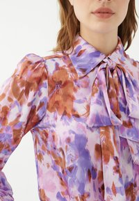 Bluse mit floralem Muster in Lila, Braun und Pink, mit strukturiertem Kragen und Schleifen-Applikation, aus leichtem Stoff gefertigt.