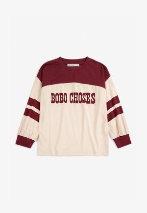 Enfant portant un t-shirt à manches longues crème et bordeaux avec le texte "BOBO CHOSE" et un jean bleu avec de grandes pièces "B" et "C", marchant vers la gauche.