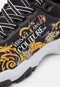 Gros plan d'une basket noire avec des lacets blancs et noirs, affichant le logo Versace Jeans Couture et des motifs baroques jaunes sur le côté.