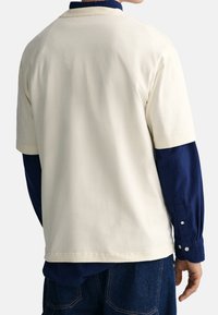  camiseta de manga corta de color crema con dobladillo cosido y fit relajado, sobre una camisa de manga larga azul marino. Textura de algodón suave.