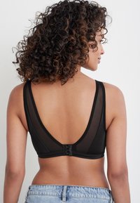 Zwarte mesh bralette met een diepe V-rug, verstelbare bandjes en een subtiele haak-en-oogsluiting aan de achterkant. Spijkershorts zichtbaar.