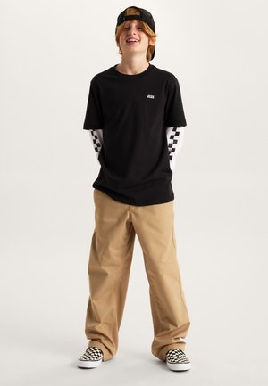 Maglietta nera a maniche corte con il logo Vans, indossata sopra una camicia bianca a maniche lunghe con motivo a scacchi nero. Pantaloni beige larghi e scarpe slip-on a scacchi.