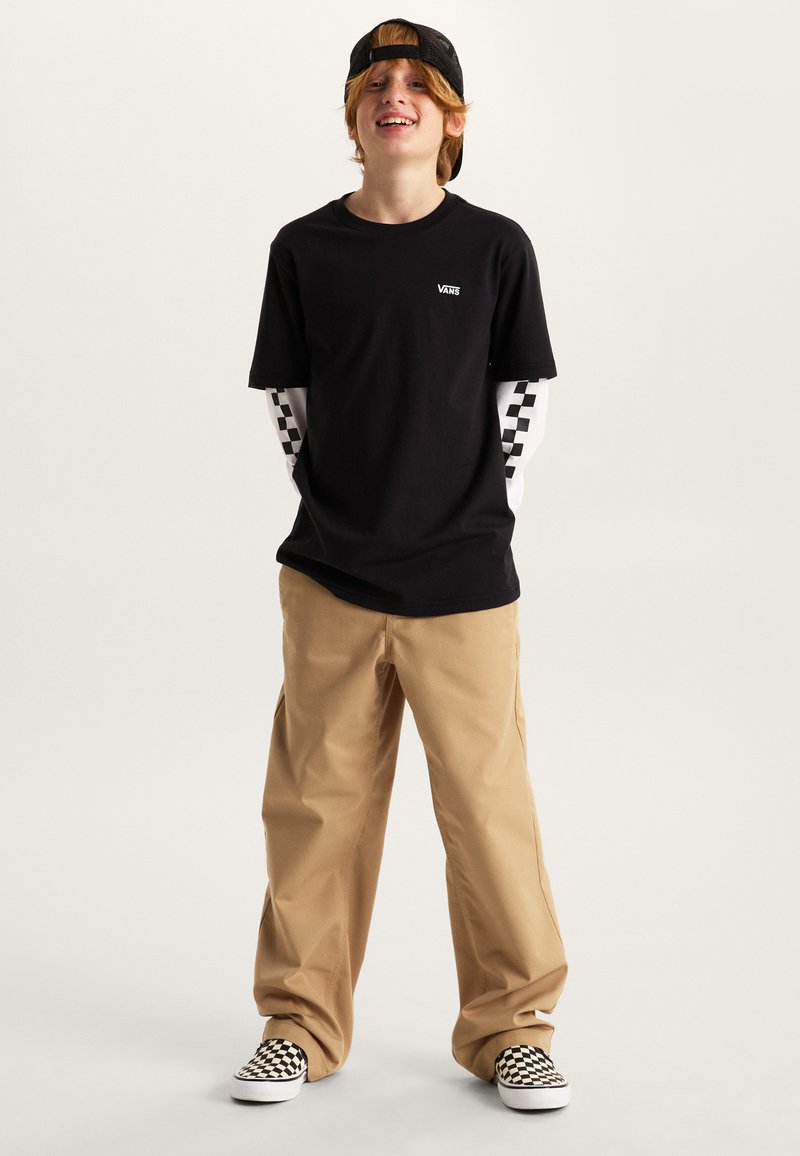 Czarny t-shirt z krótkim rękawem z logo Vans, noszony na białej bluzce z długim rękawem z czarno-białym wzorem w kratę. Beżowe, luźne spodnie i wsuwane buty w kratę.