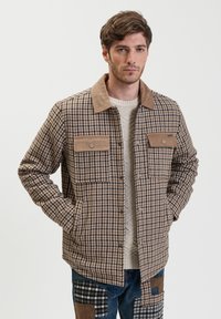Bruin en tan geruite jas met een fluwelen kraag, twee voorkezen en een knoopsluiting, gedragen over een lichte trui en blauwe spijkerbroek.
