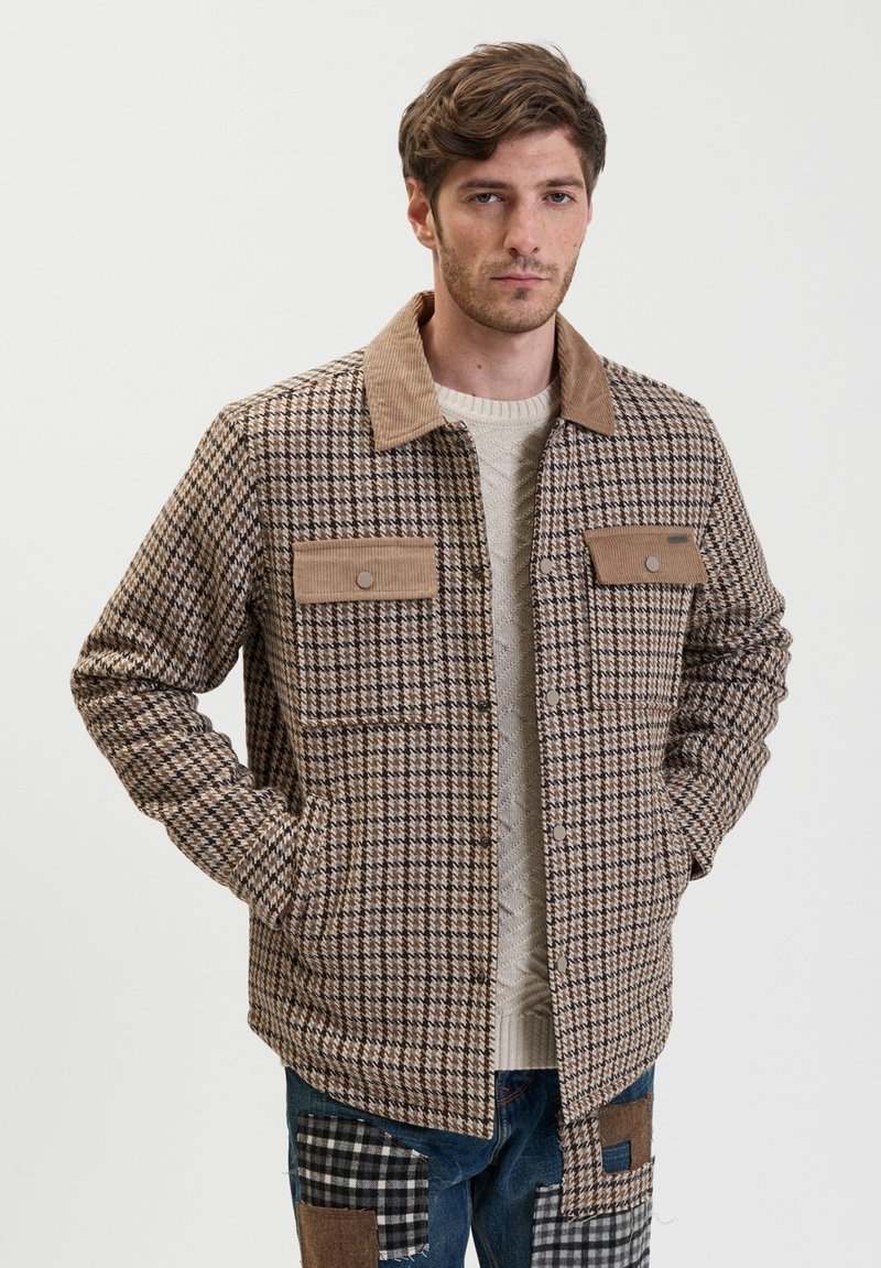 Bruin en tan geruite jas met een fluwelen kraag, twee voorkezen en een knoopsluiting, gedragen over een lichte trui en blauwe spijkerbroek.