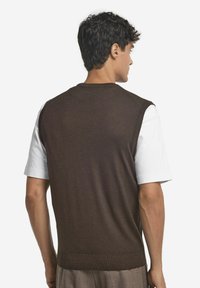 Brun ärmlös tröja med texturerad finish, bärs över en vit skjorta. Har en rund hals och ribbat avslut. Enkel och modern design.