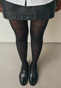Čierne vzorované pančuchy s mriežkovaným dizajnom, skombinované s čiernymi poltopánkami. Textúra pančúch je polopriehľadná, čo zdôrazňuje vzor.