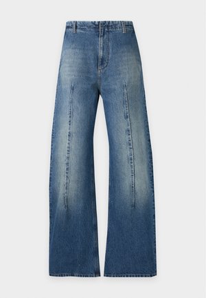 Femme debout, portant un T-shirt noir avec le texte "Margiela", un jean bleu à jambes larges, des chaussures noires, tenant un petit sac à main noir, fond blanc.