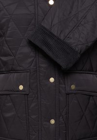 Veste matelassée noire avec poignets côtelés, deux poches avant à rabat et boutons-pression dorés au centre.