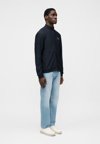 Μπλε ναυτικό μπουφάν με φερμουάρ και υφασμένη υφή, πλαϊνές τσέπες και μικρό λογότυπο. Συνδυάζεται με ανοιχτό μπλε τζιν παντελόνι με ίσια γραμμή και λευκά αθλητικά παπούτσια.