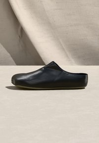 Μαύρο δερμάτινο mule παπούτσι χωρίς κορδόνια, με στρογγυλεμένη μύτη και επίπεδη σόλα, εμφανίζεται σε ουδέτερο φόντο με απαλές σκιές.