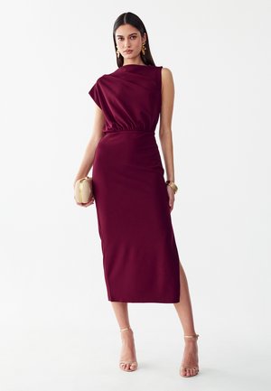 Mujer con vestido midi burdeos sin mangas con abertura lateral, pendientes dorados, pulseras, sosteniendo un clutch pequeño beige y con tacones dorados.