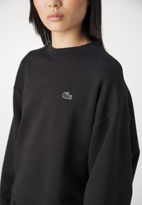 Femme portant un sweat-shirt noir avec un petit logo crocodile à contour blanc sur la poitrine, sur un fond uni.