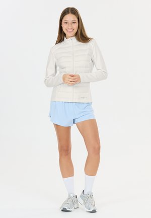 Jonge vrouw met licht jack, blauwe korte broek, witte sokken en grijze sneakers, staand en glimlachend tegen een witte achtergrond.