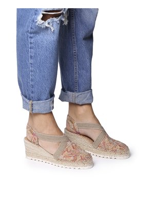 Stopy w beżowych espadrylach na koturnie z kwiatowym wzorem i z paskami na kostce, w połączeniu z podwiniętymi, podartymi niebieskimi dżinsami na białym tle.