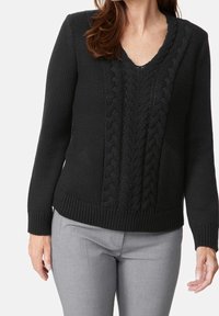 Svart stickad tröja med V-ringning, med ett kabelstickat mönster på framsidan och ribbade ärmslut och nederkant. Bärs med grå byxor.