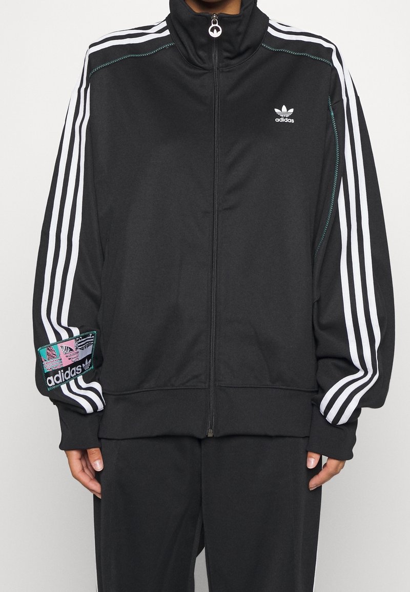 Musta vetoketjullinen verkkaritakki korkealla kauluksella, valkoiset sivuraidat ja monivärinen logopatch vasemmassa hihassa. Valmistettu sileästä kankaasta.