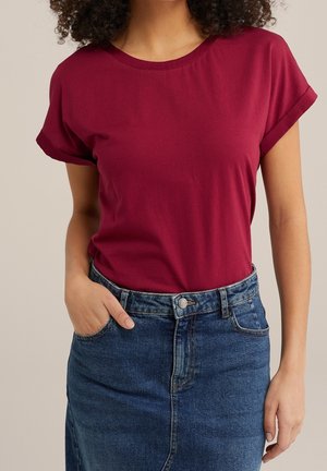 Femme portant une chemise bordeaux à manches courtes rentrée dans une jupe en jean bleue, une main dans la poche, sur un fond neutre.