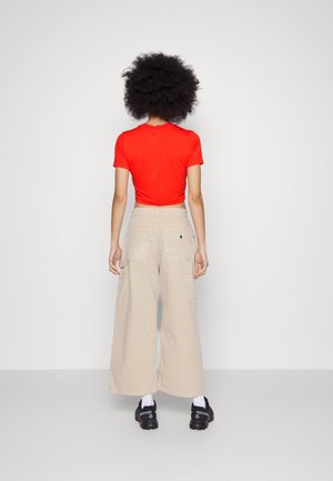 Osoba o dużych, kręconych włosach stoi twarzą do białej ściany, ubrana w czerwoną bluzkę z krótkim rękawem, beżowe spodnie o szerokich nogawkach, białe skarpetki i czarne buty.