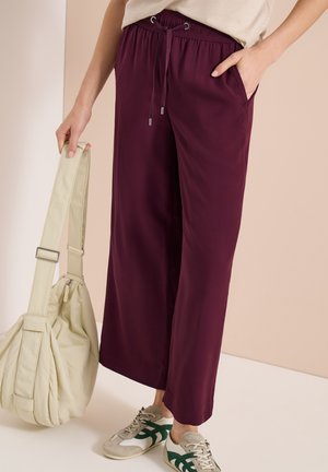 Persona que lleva pantalones sueltos color burdeos con cintura elástica con cordón, zapatillas beige y verdes, sosteniendo una gran bolsa beige por la correa.