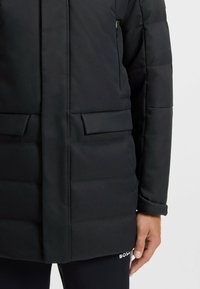Black padded coat with a straight cut, featuring side pockets and a zipper. Made from textured fabric, showcasing a simple design.
