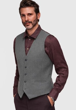 Mężczyzna z krótkimi włosami i brodą, ubrany w szarą kamizelkę zapinaną na guziki na ciemnofioletowej koszuli, ręce w kieszeniach, stoi na jednolitym tle.