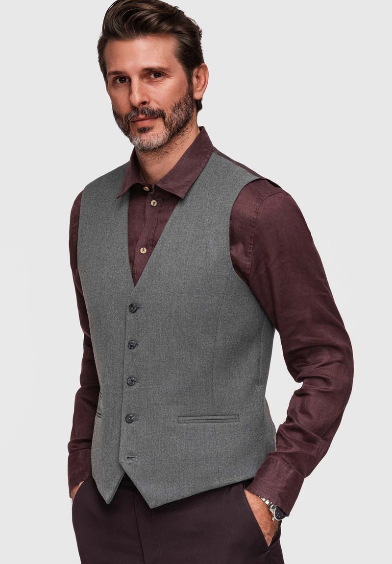 Homme aux cheveux courts et à la barbe portant un gilet gris boutonné sur une chemise violet foncé, mains dans les poches, debout devant un fond uni.