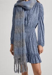 Sininen ja harmaa gradientti huivi, joka on valmistettu pehmeästä, teksturoituneesta kankaasta. Varustettu fringed-reunoilla ja hienovaraisella logo-tarralla yksityiskohtana.