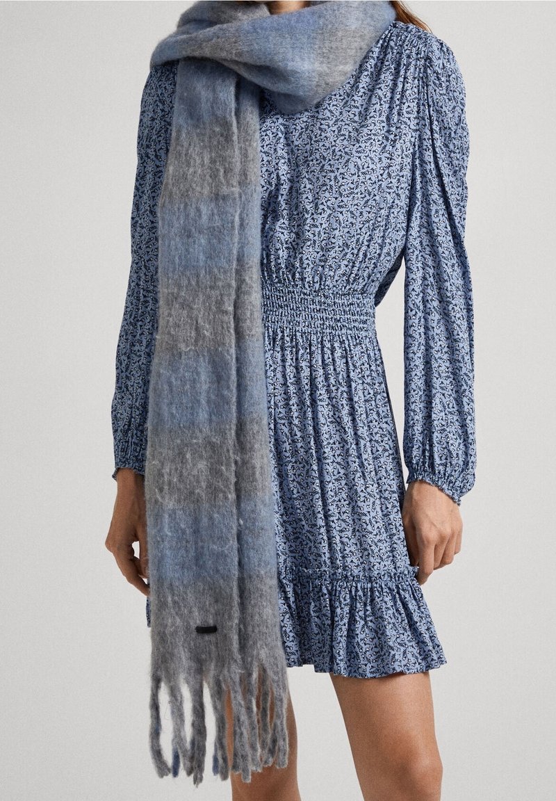 Sininen ja harmaa gradientti huivi, joka on valmistettu pehmeästä, teksturoituneesta kankaasta. Varustettu fringed-reunoilla ja hienovaraisella logo-tarralla yksityiskohtana.