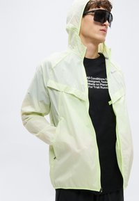 Ελαφρύ, ημιδιαφανές ζακέτα σε πράσινο λάιμ με κουκούλα, κλείσιμο με φερμουάρ και λεπτομέρειες από δίχτυ. Φοριέται πάνω από μαύρο μπλουζάκι με λευκό κείμενο.