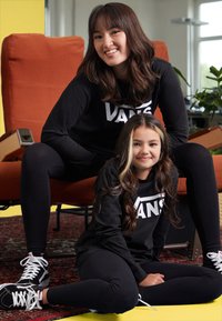To personer iført sorte langærmede trøjer med et stort "VANS" logo og sorte leggings. Begge har sorte og hvide sneakers på. En orange stol i baggrunden.