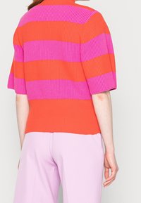 Ribbad stickad topp i orange och rosa horisontella ränder, med korta ärmar och hög hals, kombinerad med ljusrosa vida byxor.