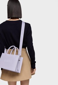 Borsa tote lilla chiaro con logo "TOUS" in rilievo, completata da una tracolla di tessuto con testo "TOUS" ripetuto. Texture liscia e design strutturato.