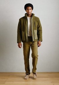 Olivgrüne Fleecejacke mit Reißverschluss und zwei Vordertaschen, kombiniert mit beigen Hosen und hellbraunen Schuhen, vor einem Holzboden-Hintergrund.