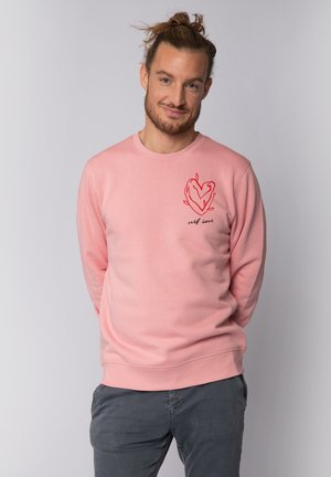 Sweatshirt rose clair à manches longues, avec un motif de cœur rouge et le texte "amour de soi" sur la partie supérieure gauche de la poitrine. Texture de tissu douce.