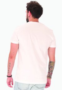 Uomo con capelli corti e scuri, tatuaggi su entrambi i avambracci, che indossa una t-shirt bianca semplice e jeans azzurri chiari, di spalle rispetto alla fotocamera.