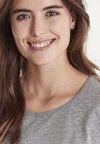 Portræt af en smilende person med langt, bølget brunt hår. Iført en grå kortærmet skjorte med rund halsudskæring. Klar hud og diskret smykker.