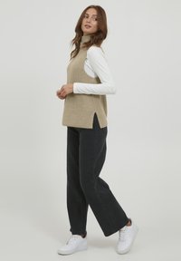 Pull en maille beige sans manches avec un col montant et des fentes latérales, associé à un pantalon noir à jambes larges et des baskets blanches.