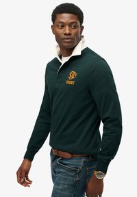 Polo de rugby verde oscuro con cuello blanco, que presenta un logotipo bordado naranja y el texto "RUGBY". Se lleva con jeans azules y un cinturón marrón.