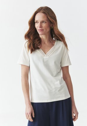 T-shirt blanc à manches courtes avec un col en V texturé, fabriqué dans un matériau doux, porté avec un pantalon large bleu foncé.